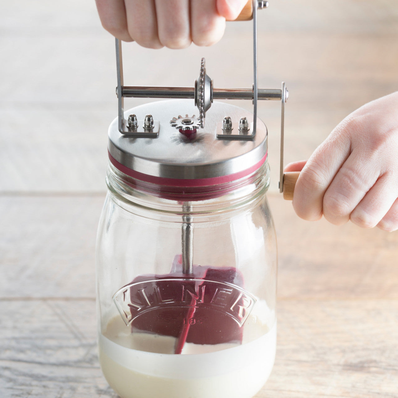 Eine Hand dreht eine Kurbel an einem Glas-Butterrührer mit Sahne und kreiert selbstgemachte Butter für eine stilvolle Küchenergänzung.