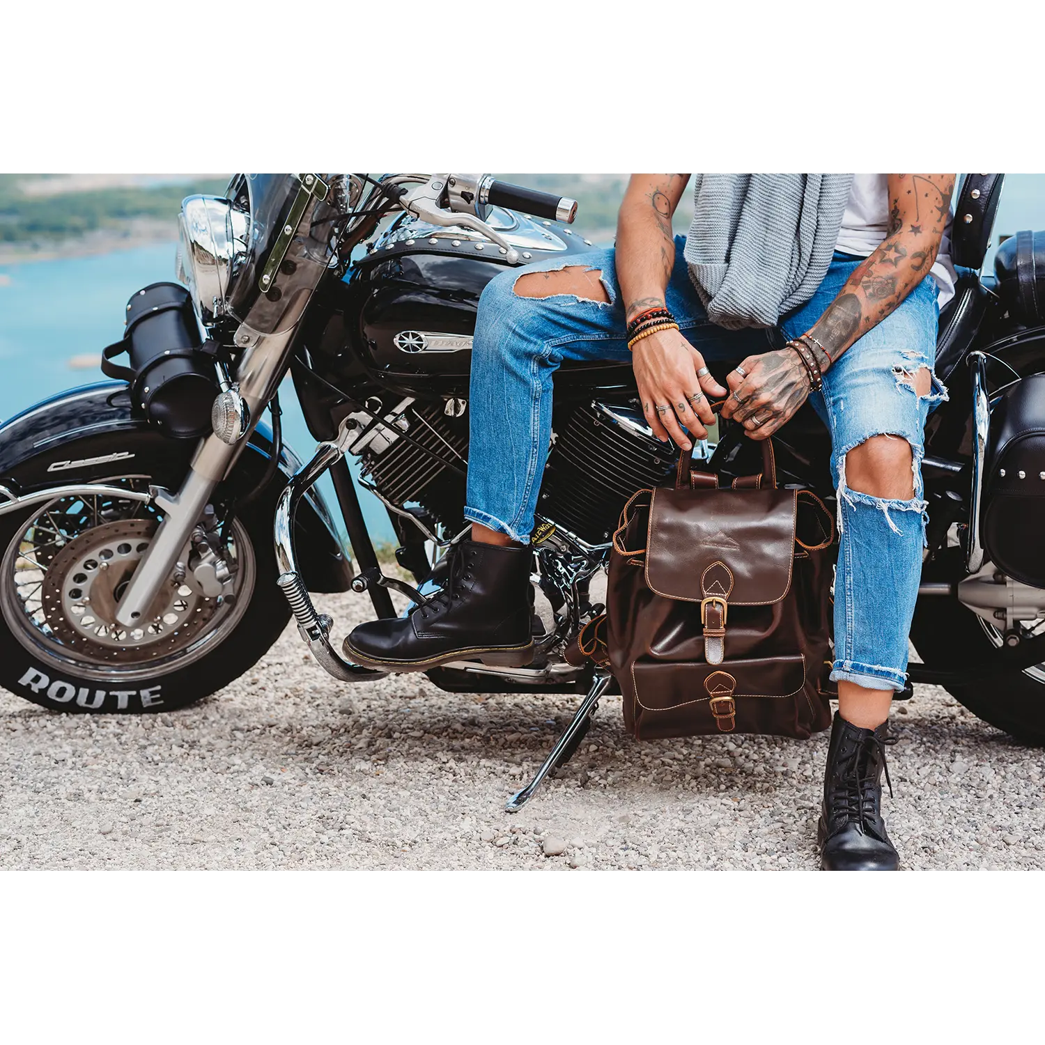 An einem malerischen Gewässer sitzt jemand mit tätowierten Armen auf einem Motorrad. Er trägt zerrissene Jeans, einen Schal und schwarze Stiefel. Daneben steht ein Vintage-Tagesrucksack: der braune Leder-Rucksack JACOB'S WAY, der perfekt zu seinem abenteuerlichen Reisebegleiter-Geist passt.