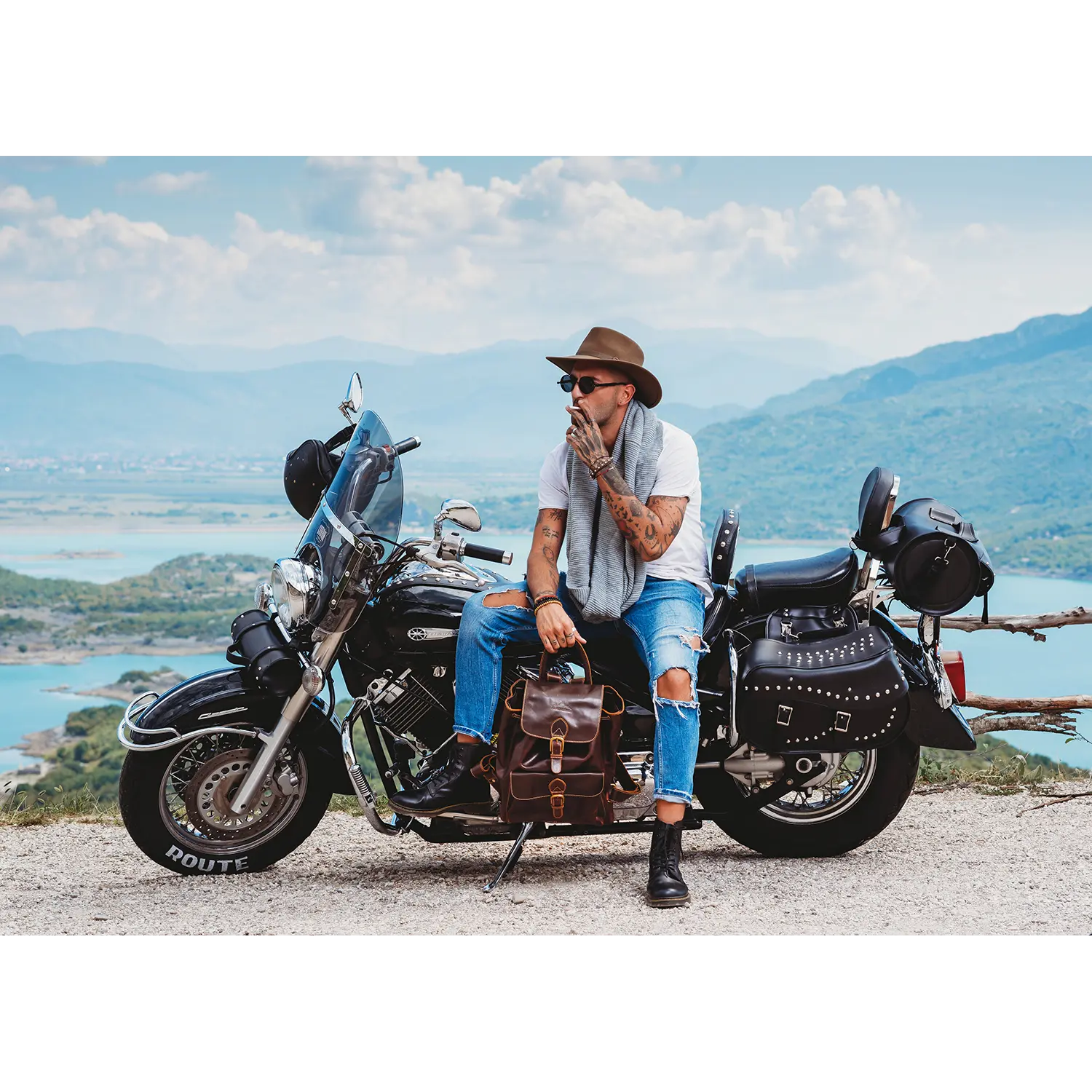 Eine Person mit Hut und Sonnenbrille sitzt nachdenklich auf einem Motorrad an einem Aussichtspunkt. Neben ihr lehnt der Leder-Rucksack JACOB'S WAY am Sitz. Berge, ein See und leicht bewölkter Himmel heben ihr graues Oberteil und die zerrissenen Jeans wunderschön hervor.