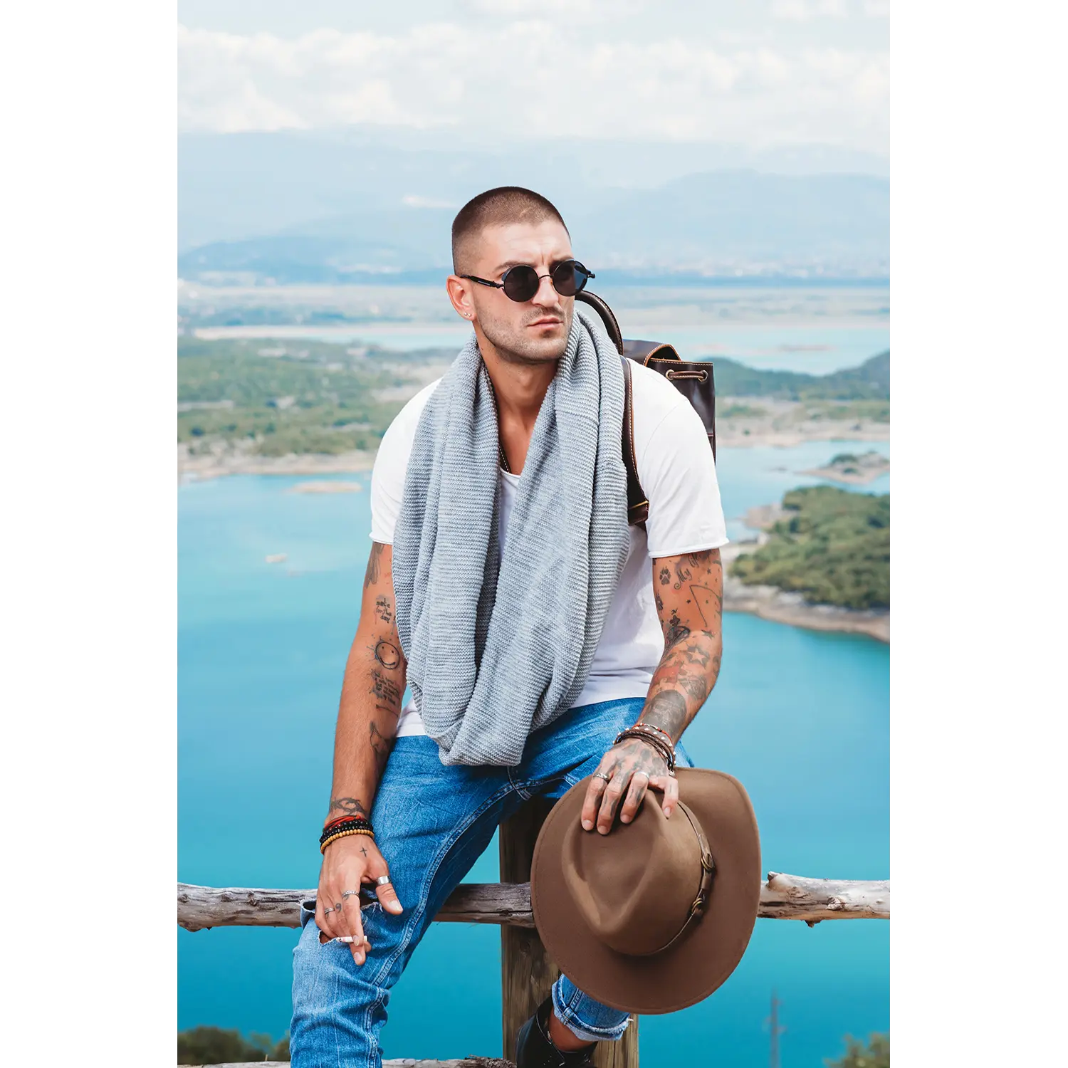 Ein Mann mit Sonnenbrille, Tattoos und grauem Schal sitzt auf einem Holzgeländer mit Blick auf blaues Wasser und Berge. Neben ihm steht der Lederrucksack JACOB'S WAY. Er hält einen braunen Hut in der Hand und trägt ein weißes T-Shirt und Jeans unter einem leicht bewölkten Himmel.