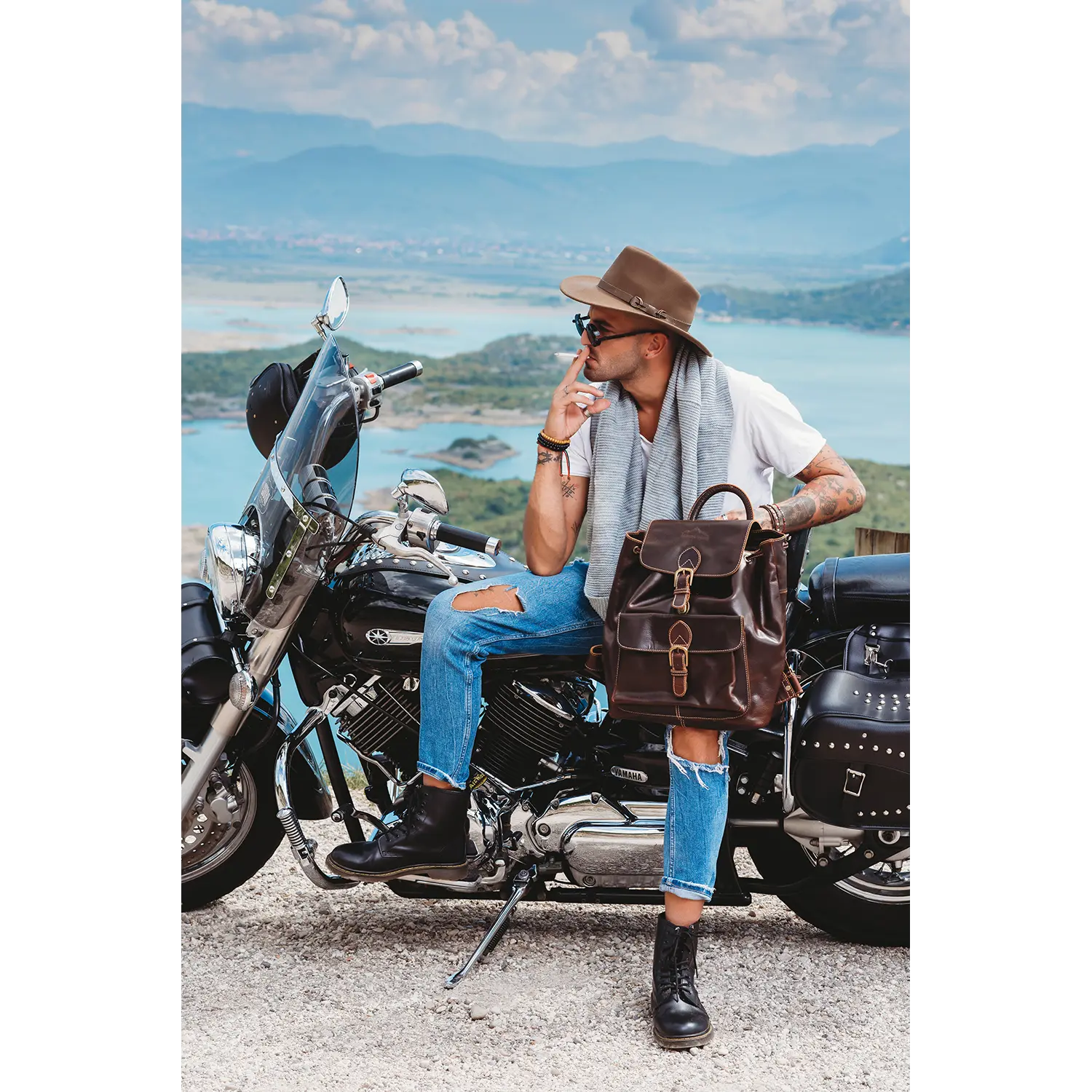 Ein Mann mit Hut und Sonnenbrille sitzt auf einem Motorrad an einem malerischen See, umgeben von Bergen. Er trägt ein weißes Hemd, zerrissene Jeans und Stiefel und hält den Leder-Rucksack JACOB'S WAY in der Hand – eine perfekte Kombination aus Stil und Funktionalität für seinen Abenteuergeist.