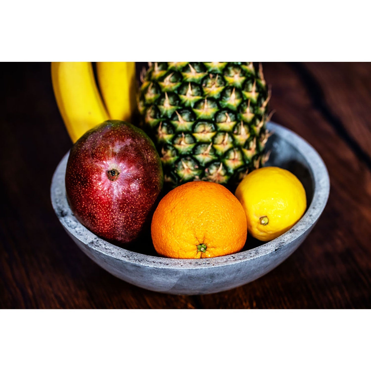 Eine handgefertigte, ästhetische große runde Betonschale enthält eine bunte Auswahl an Früchten auf einer Holzoberfläche, darunter eine Ananas, Bananen, eine Mango, eine Orange und eine Zitrone.