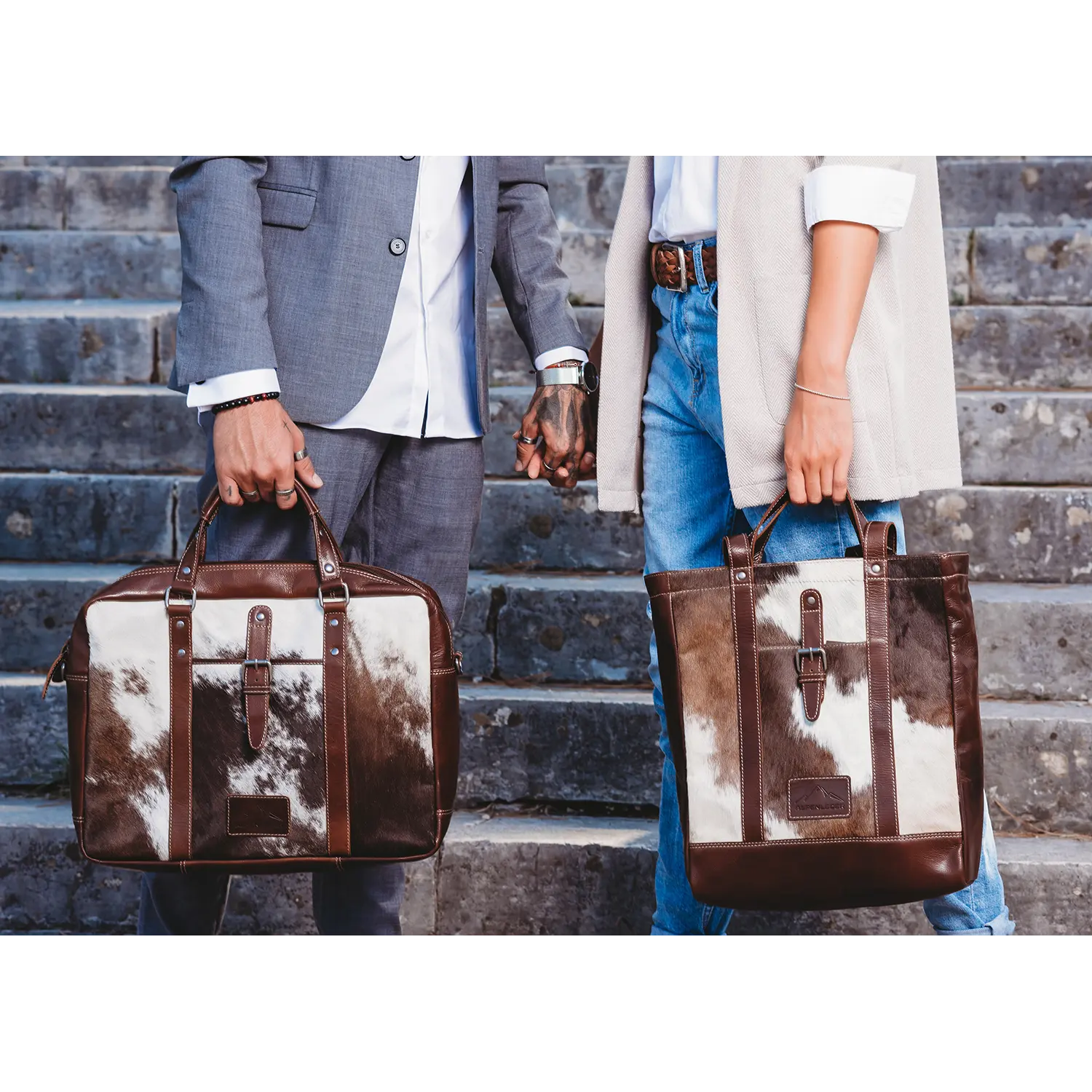 Ein Paar hält Händchen auf Steinstufen und trägt Alpenleder Taschen; er trägt einen grauen Anzug, sie ein weißes Hemd, Jeans und eine beige Strickjacke. Ihr Look wird durch das Rindsledermuster ihrer LACKL Aktentaschen aufgewertet.