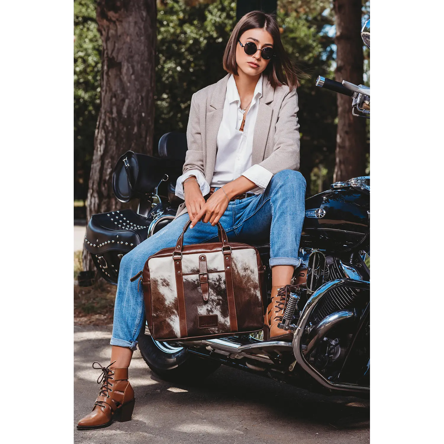 Eine Person in beigem Blazer, weißem Hemd und Jeans sitzt auf einem Motorrad und hält eine stylische Aktentasche von LACKL in der Hand. Sie trägt eine Sonnenbrille und braune Stiefeletten. Bäume im Hintergrund verleihen dieser Outdoor-Szene ein einzigartiges Flair – ein echtes Unikat.