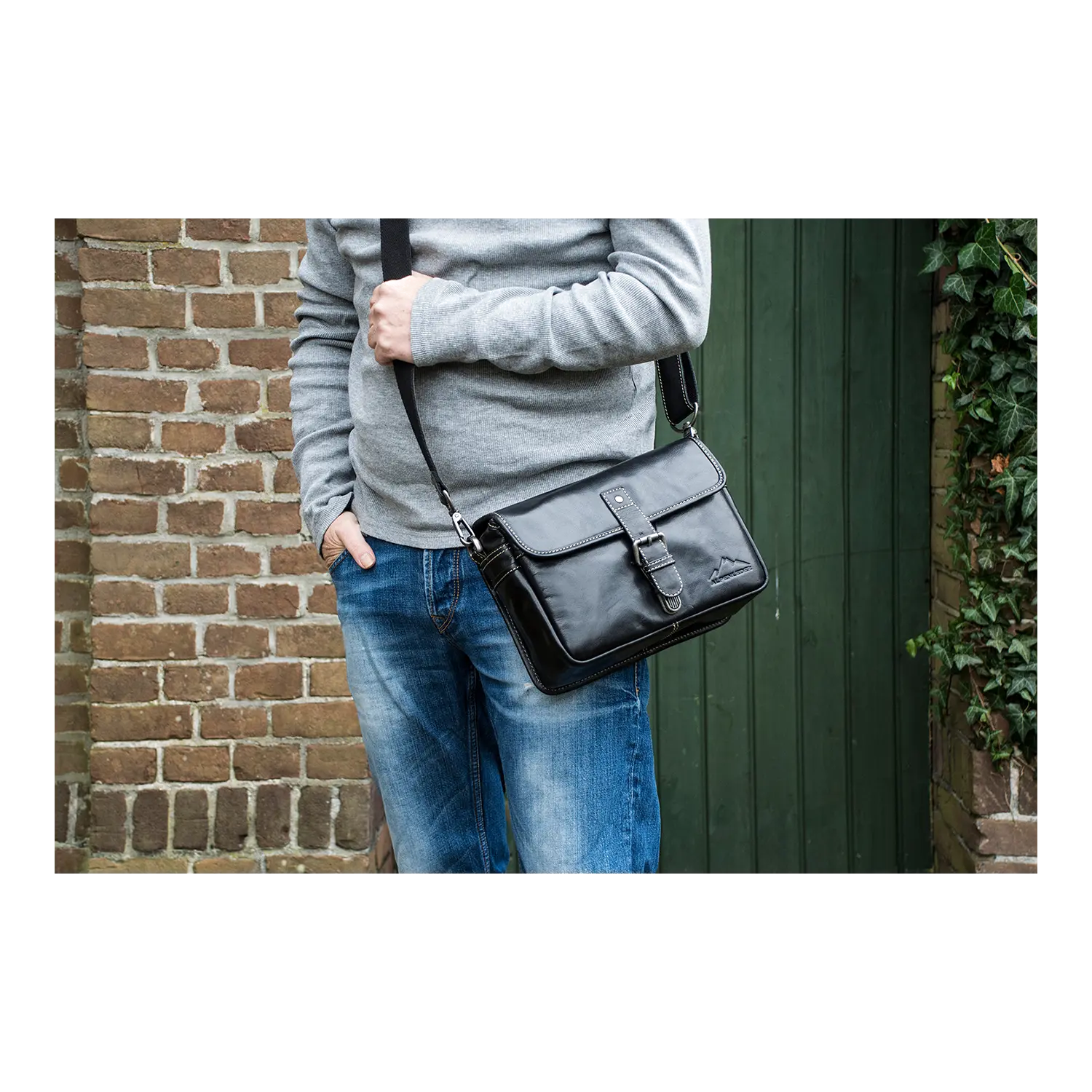 Eine Person in einem grauen Pullover und blauen Jeans steht neben einer Backsteinmauer und einer grünen Holztür und hält eine Alpenleder-Kameratasche PRESSE aus schwarzem Leder mit Schnalle in der Hand.