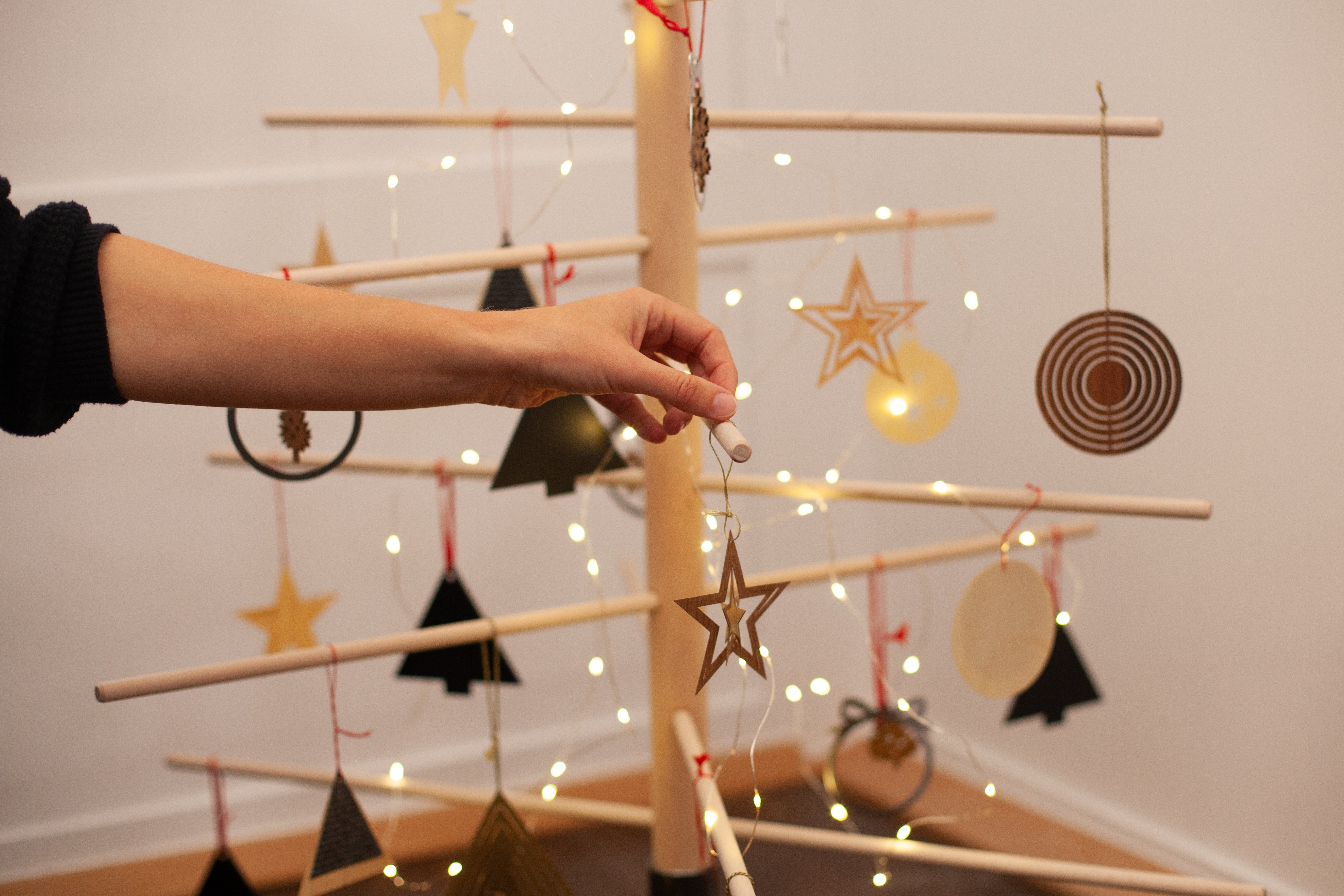 Eine Hand hängt ein Sternornament an einen minimalistischen Buchenholz-Weihnachtsbaum mit Lichtern und geometrischen Dekorationen und fängt den Charme nachhaltiger Weihnachtsbäume ein.