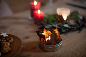Ein festlicher Teelichthalter mit Hirsch- und Baumsilhouetten leuchtet auf einem Nussbaumholztisch, umgeben von Keksen und Kerzen.