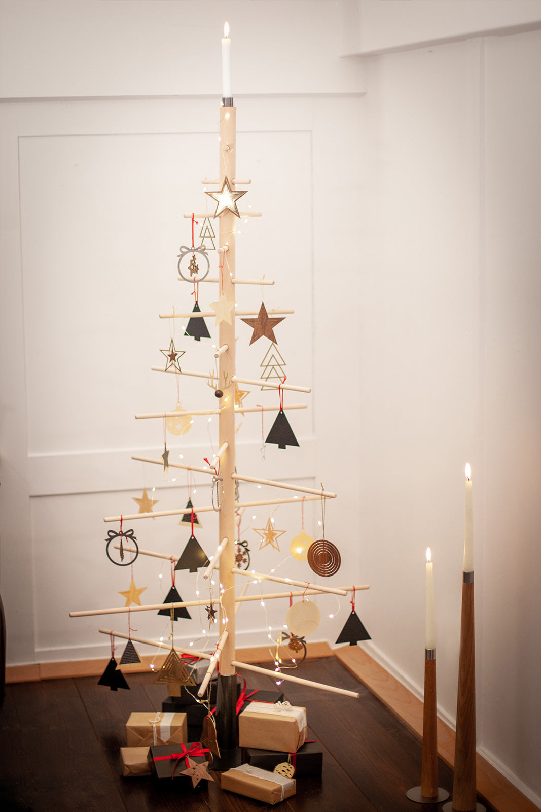 Minimalistischer abstrakter Baum aus Buchenholz mit Ornamenten, Lichterketten und Geschenken steht neben hohen, leuchtenden Kerzen – eine stilvolle Wahl für nachhaltige Weihnachtsbäume im modernen Raum.