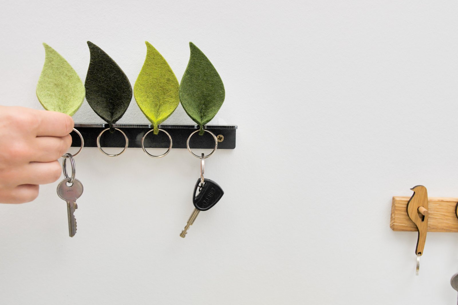 Eine Hand hängt Schlüssel an ein blattförmiges Schlüsselbrett, das von einem Designer aus Naturfilz gefertigt und an einer weißen Wand montiert wurde.