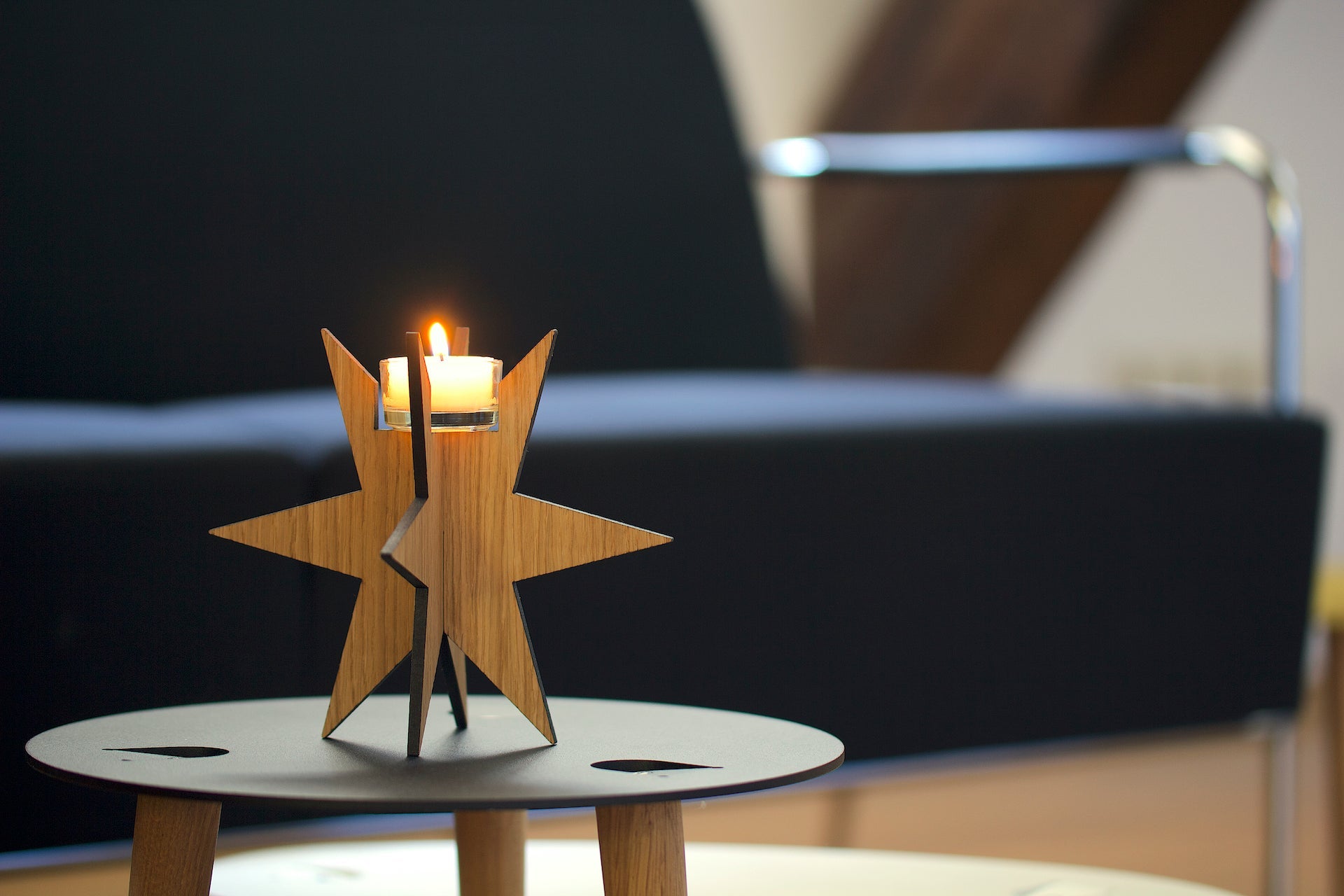 Eine brennende Kerze in einem Teelichthalter „Sternenlicht“ aus Eichenholz steht auf einem runden Tisch vor einem dunkelblauen Sofa und verleiht der Umgebung Schwarzwald-Charme.