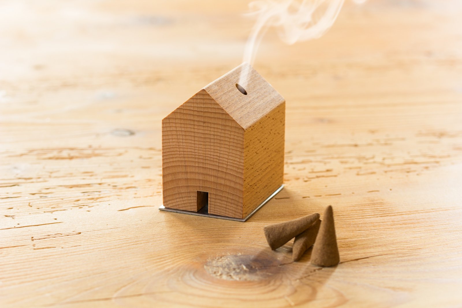 Räucherstäbchenhalter aus Holz in Form eines Hauses, inspiriert von Schwarzwaldhöfen und entworfen von Jan Hartmann, mit aufsteigendem Rauch neben drei braunen Räucherkegeln auf einem Holztisch.