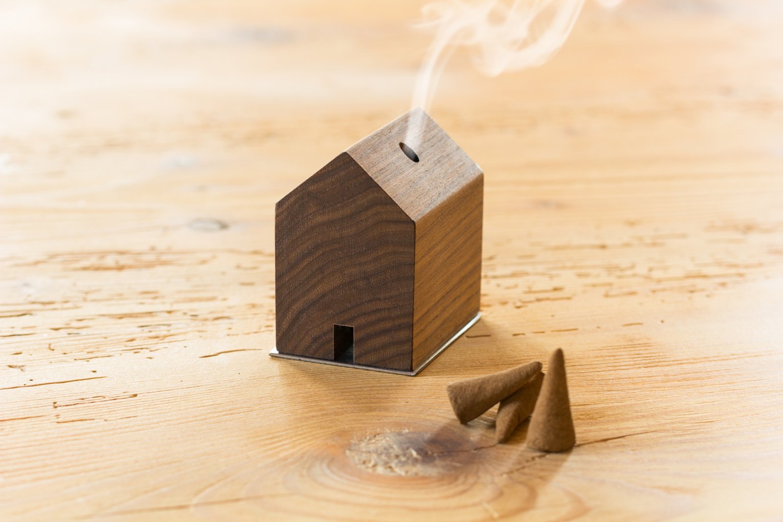 Ein hölzerner Räucherbrenner „RÄUCHERMÄNNCHEN RAUCHHAUS“, inspiriert von Schwarzwaldhöfen, hat die Form eines Hauses und stößt Rauch aus, während Räucherkegel in der Nähe auf einer Holzoberfläche ruhen.