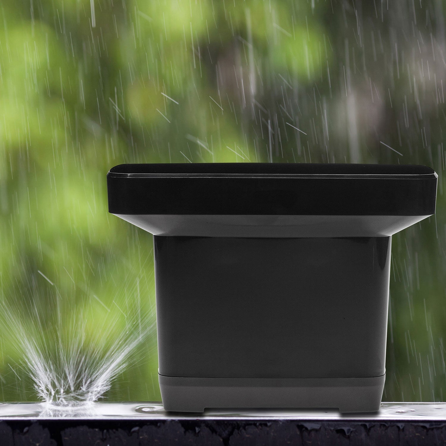 Ein schwarzer, rechteckiger Blumentopf mit grauem Sockel steht auf einer regennassen Oberfläche und reflektiert die Messwerte des Digitalen Funk-Regenmessers. Spritzendes Wasser verstärkt die Unschärfe der grünen Natur im Hintergrund und sorgt für eine ruhige Szenerie.