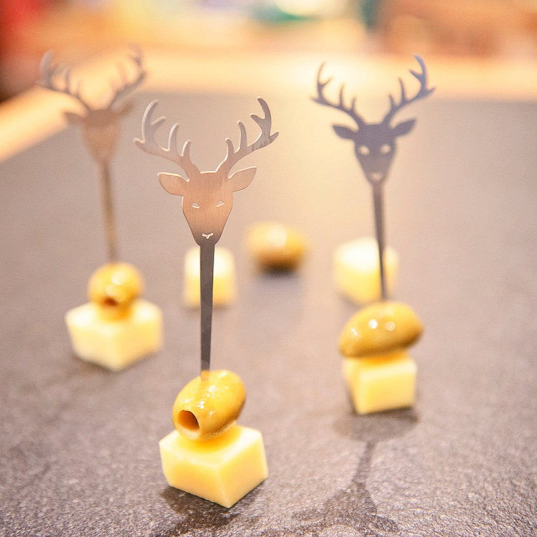 Käsewürfel und grüne Oliven auf Zahnstochern mit Edelstahl-Hirschkopf-Motiven verleihen Ihrer Küche eine stilvolle Note, hübsch arrangiert auf einer dunklen Oberfläche – perfekt als Partysticker-Highlights für jede Zusammenkunft.