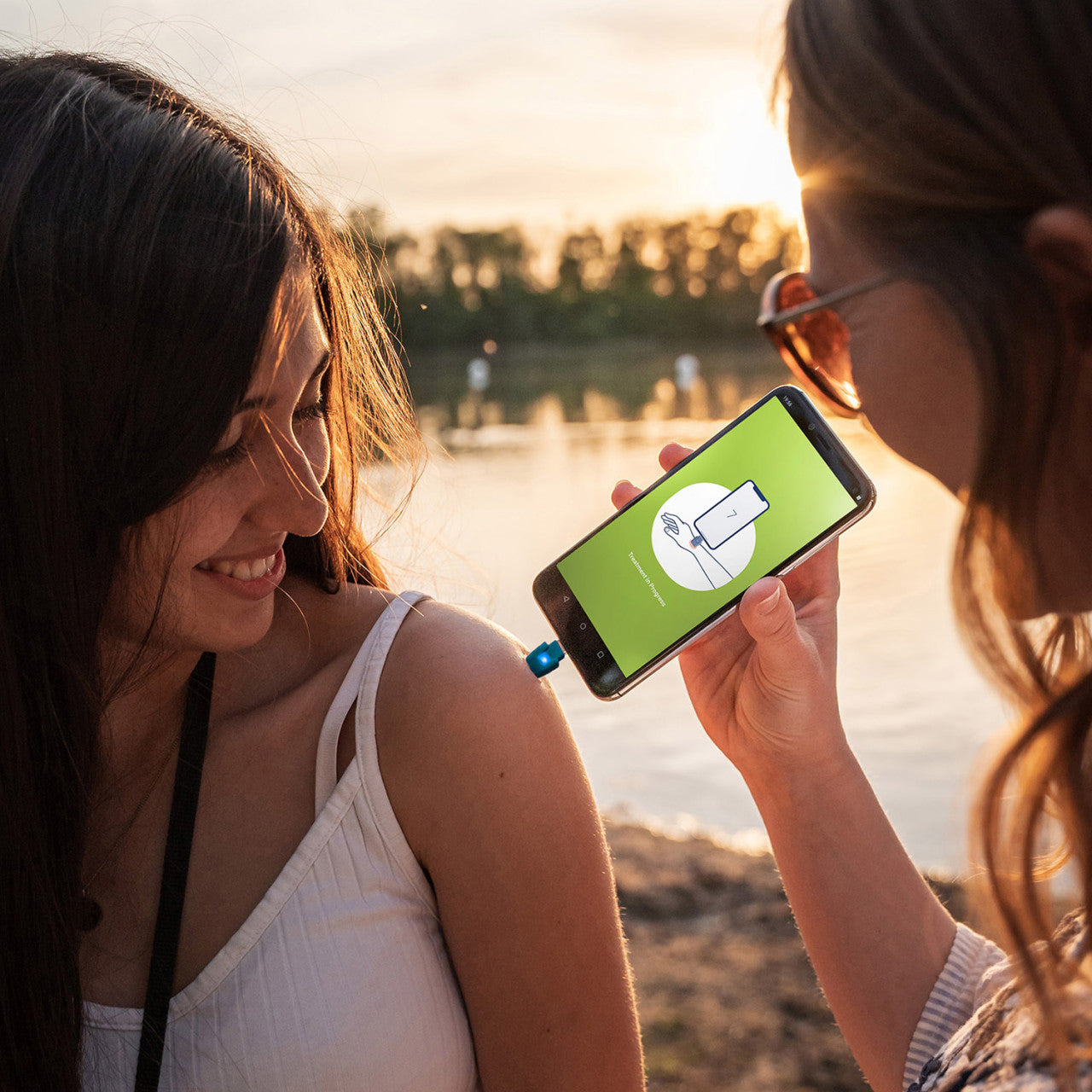 Zwei Frauen an einem See. Eine scannt den Arm der anderen mit einem Smartphone, auf dem die grüne Stichheiler-App zur schnellen Insektenstichbehandlung angezeigt wird.