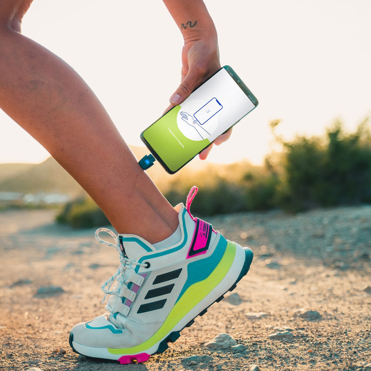 Eine Person in bunten Turnschuhen verwendet im Freien ein Smartphone mit einem tragbaren Gerät und steuert wahrscheinlich die Insektenstichbehandlung über die heat it® Stichheiler App.