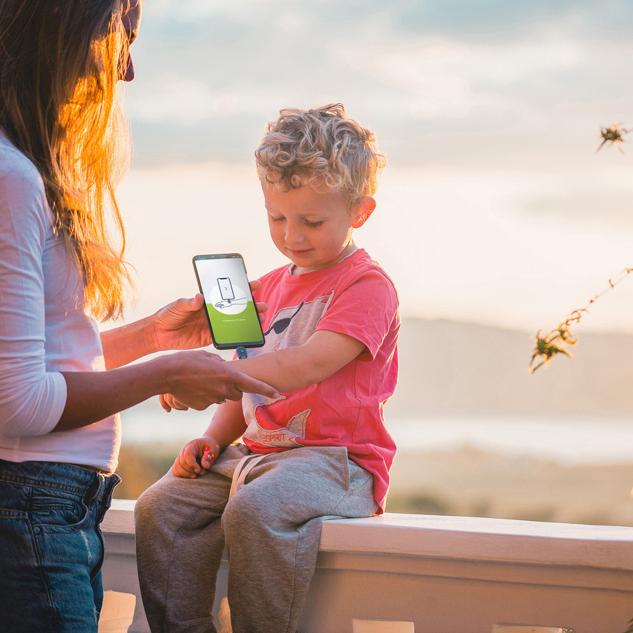 Eine Frau scannt bei Sonnenuntergang im Freien mit der heat it® Stichheiler App auf ihrem Smartphone den Arm eines Jungen zur Insektenstichbehandlung; der Junge blickt auf seinen Arm hinunter.