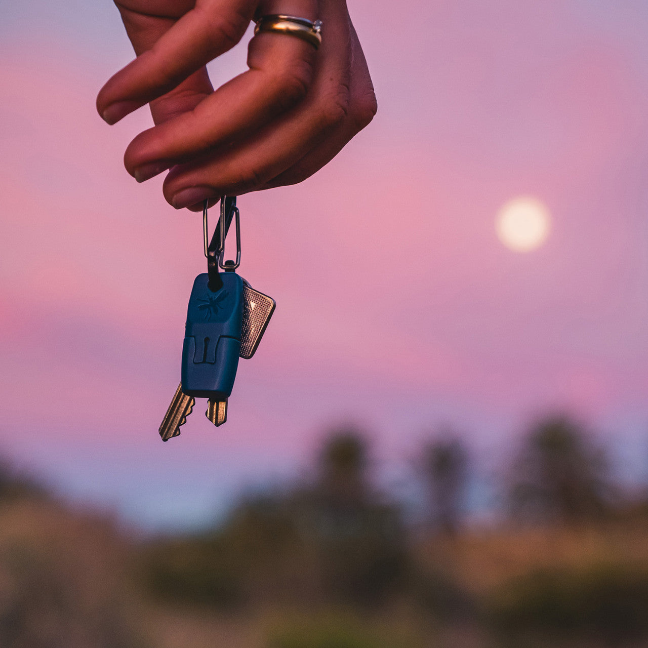 Eine Hand, die vor einem Sonnenuntergangshimmel mit einem verschwommenen Vollmond im Hintergrund den Hausschlüssel hält, erinnert an die Suche nach Trost nach einem Abenteuer – genau wie an die Entdeckung der wohltuenden Wärme der it® Insektenstich-Behandlung.
