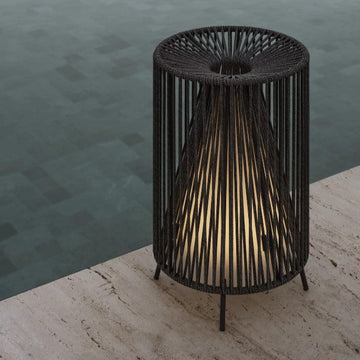 Eine moderne ROPE-Leuchte aus schwarzem Metall mit gewebtem Design, die warm am gefliesten Poolrand auf einer Steinoberfläche leuchtet – eine atemberaubende kabellose Leuchte vom Designstudio kaschkasch.
