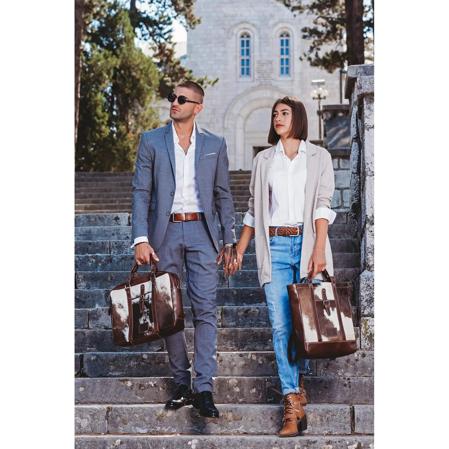 Ein elegantes Paar steigt Hand in Hand eine Steintreppe hinunter. Der Mann trägt einen grauen Anzug und eine Sonnenbrille, die Frau einen hellgrauen Blazer und Jeans. Beide tragen passende Alpenleder Shopper HALLODRI Taschen aus Kuhfell, umgeben von Bäumen und einem Steingebäude.