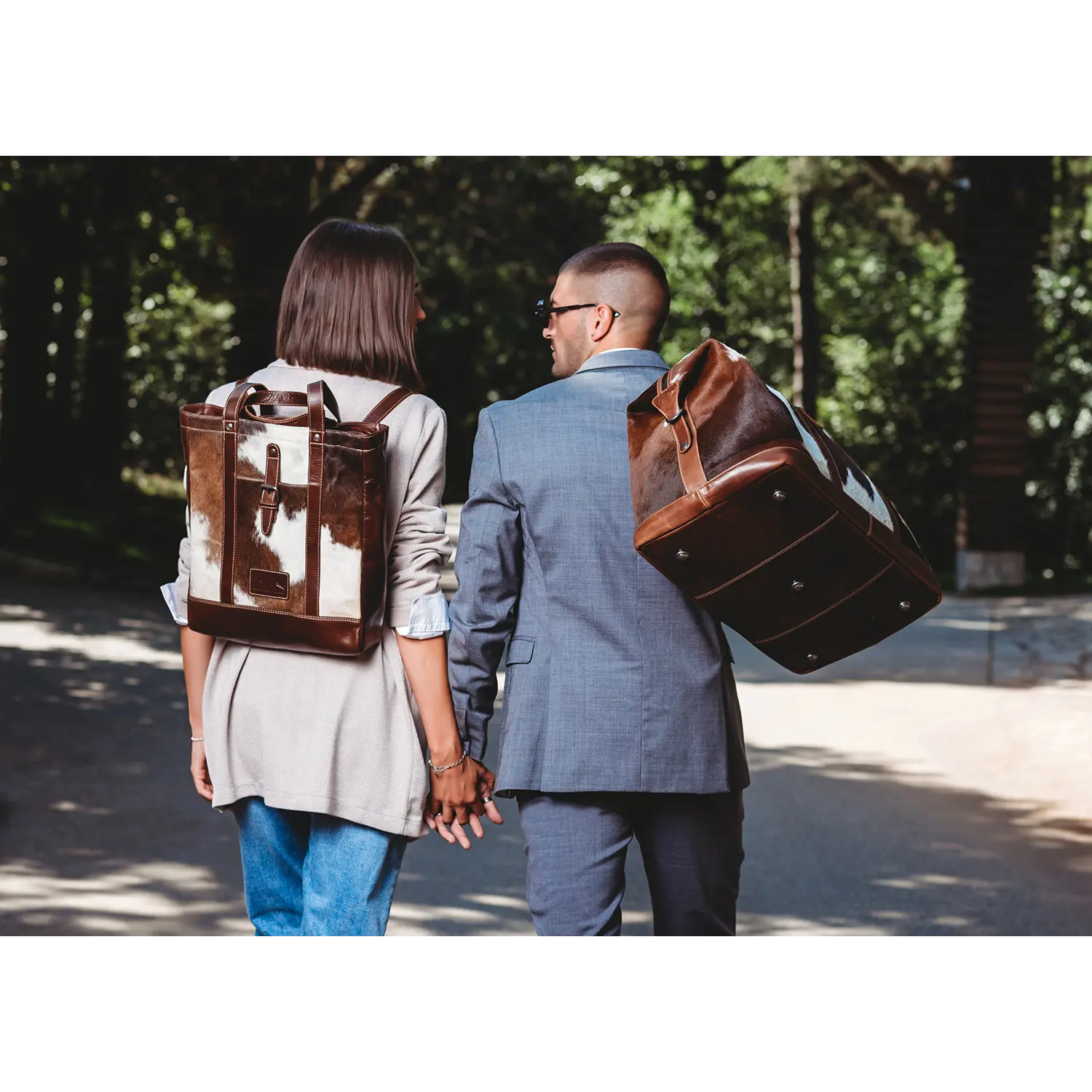 Ein Paar schlendert Hand in Hand einen sonnigen, von Bäumen gesäumten Weg entlang. Die Person links trägt Jeans, ein weißes Top und einen braunen Rucksack aus Alpenleder, während die Person rechts einen blauen Anzug trägt und eine große braune Shopper-Tasche von HALLODRI trägt.