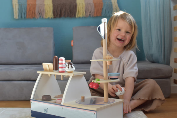Lächelndes Kind, das in einem gemütlichen Wohnzimmer mit einem hölzernen Eis-Spielset und einem magnetischen Eisportionierer auf dem Boden spielt.
