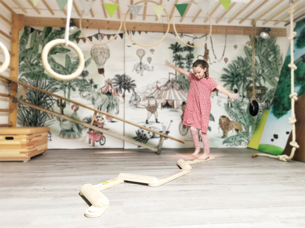 Ein Mädchen in einem roten Kleid testet ihren Gleichgewichtssinn, während sie auf Balancierstegen in einem Spielzimmer mit einem Wandgemälde zum Thema Zirkus balanciert.