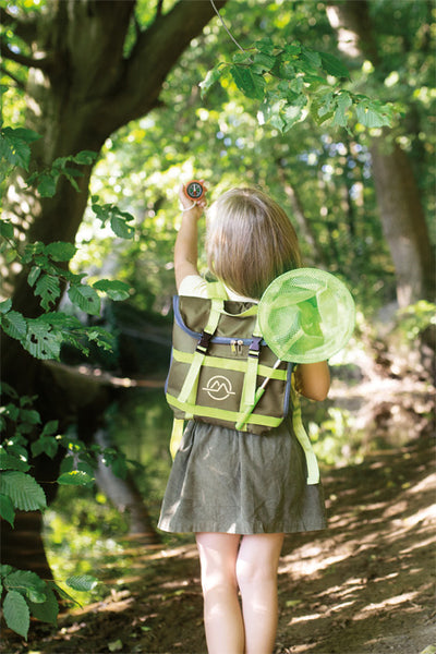 Ein kleines Kind mit Rucksack und Insektennetz erkundet einen sonnenbeschienenen Waldweg und hält einen Kompass hoch – bereit für Abenteuertouren mit seinem Lieblings-Entdeckerspielzeug.