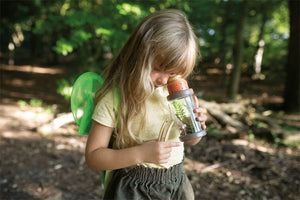 Kleines Kind untersucht im Wald eine Pflanze in einem durchsichtigen Behälter und trägt grüne Rucksackflügel – perfekt für kleine Entdecker auf Abenteuertouren.
