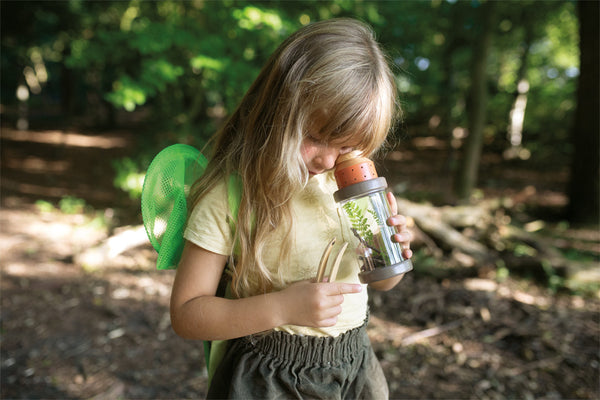 Kleines Kind untersucht im Wald eine Pflanze in einem durchsichtigen Behälter und trägt grüne Rucksackflügel – perfekt für kleine Entdecker auf Abenteuertouren.
