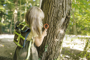 Ein kleines Kind mit Rucksack untersucht in einem sonnenbeschienenen Wald Baumrinde mit einer Lupe und macht den Moment zu einem Entdeckerspielzeug-Abenteuer.