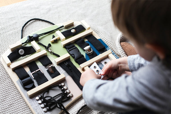Kind spielt mit einem Montessori-Lernbrett mit Schnallen, Knöpfen, Reißverschlüssen und Schnürsenkeln auf einer weichen Oberfläche – perfektes Motorikspielzeug zur Entwicklung der Feinmotorik.