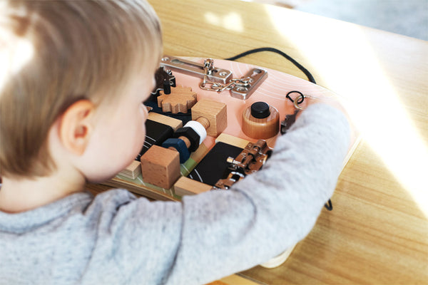 Ein kleines Kind erkundet ein Lernbrett aus Holz auf einem hellen Holztisch und unternimmt eine praktische Entdeckungsreise, inspiriert von der Montessori-Pädagogik.