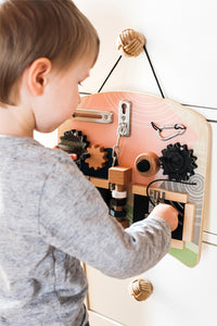 Ein Kind spielt mit einem Motorikbrett-Lernbrett aus Holz, das mit Zahnrädern, Schlössern und verschiedenen taktilen Objekten ausgestattet ist, die von der Montessori-Pädagogik inspiriert sind.
