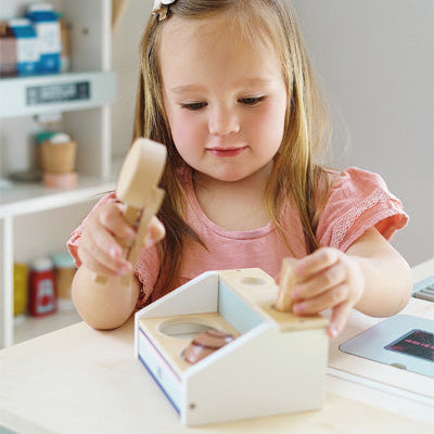 Junges Mädchen in einem rosa Hemd spielt mit einer hölzernen Spielzeugkasse in einem Spielladen, daneben eine Kinderküche und ein Eiscreme-Spielset mit magnetischem Eisportionierer.