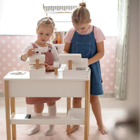 Zwei junge Mädchen spielen in einem rosa, gepunkteten Spielzimmer an einer Spielzeugkasse mit Kaufladen-Zubehör.