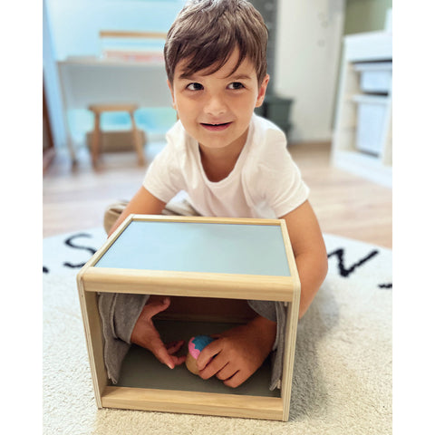 Ein kleiner Junge lächelt, während er in eine hölzerne Fühlbox greift und ein Sinneserlebnis genießt, als er darin nach einem kleinen Spielzeug tastet.