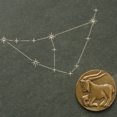 Sternbild Steinbock in Weiß gezeichnet mit einer goldenen Ziegenmünze und Sternzeichen Bronze auf dunklem Naturschiefer, darunter Steinbock geschrieben.