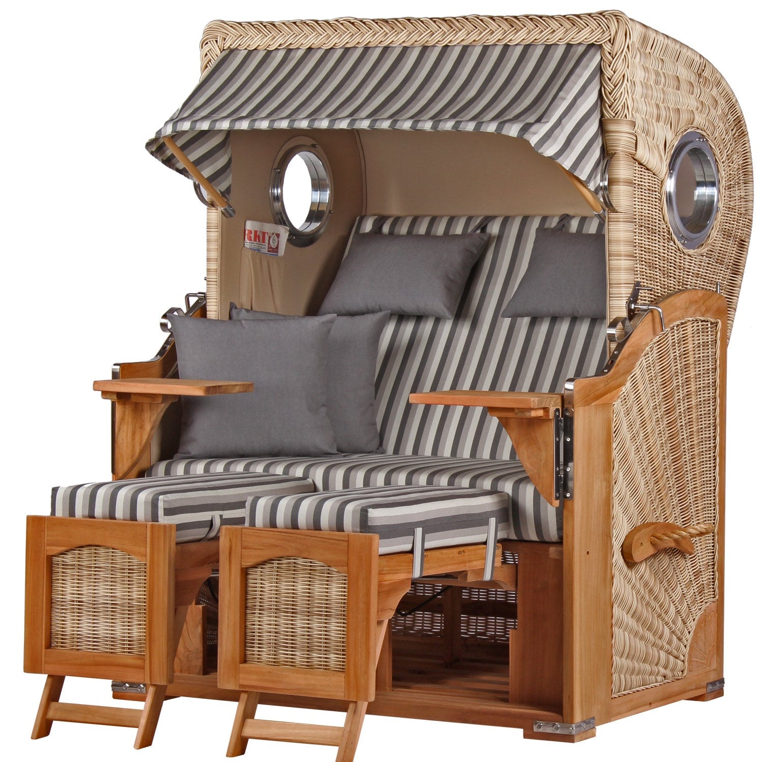 Strandkorb aus Korbgeflecht mit gestreiften Polstern, Baldachin, Kissen, Fußstützen und eleganten Armlehnen aus Teakholz – ein stilvolles Outdoor-Möbel zum Entspannen am Meer.