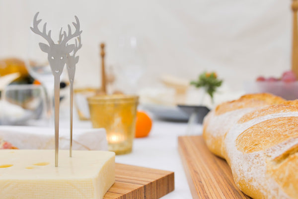 Ein Käseblock mit einem Edelstahl-Hirschspieß neben Brotlaiben auf einem Holzbrett – perfekt für Ihre nächste Party.