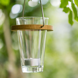 Ein gläserner Licht-Eimer mit einer kleinen Kerze hängt an einem Baumzweig, umgeben von grünen Blättern, und schafft eine stimmungsvolle Atmosphäre im Garten.