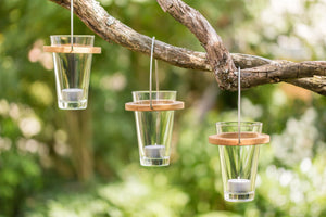 Drei gläserne Licht-Eimer-Kerzenhalter mit Holzdeckel hängen an einem Ast und sorgen für stimmungsvolle Atmosphäre im grünen Garten.